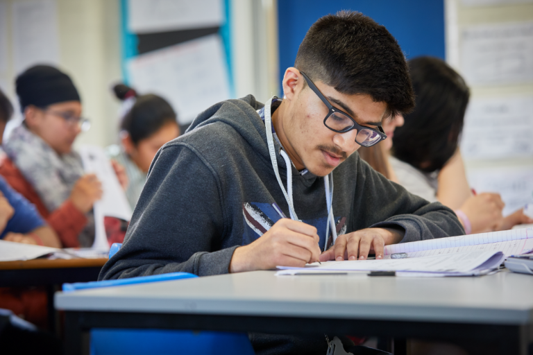 photo of male student sat at desk writing in workbook within classroom