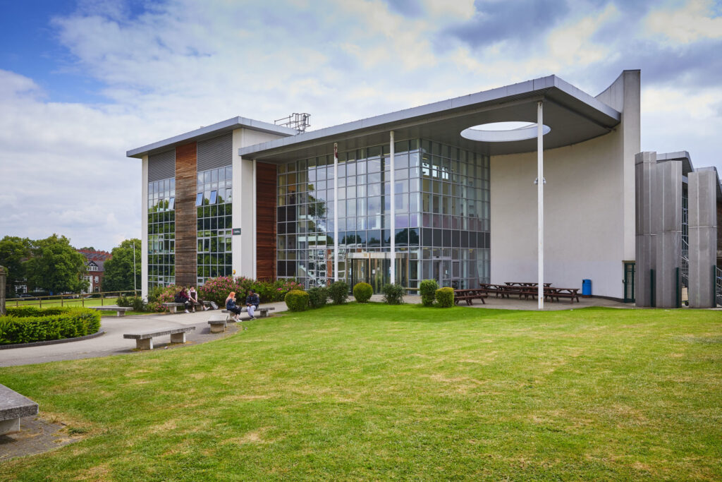 Modern glass building with people sitting outside on benches and grass under a partly cloudy sky.