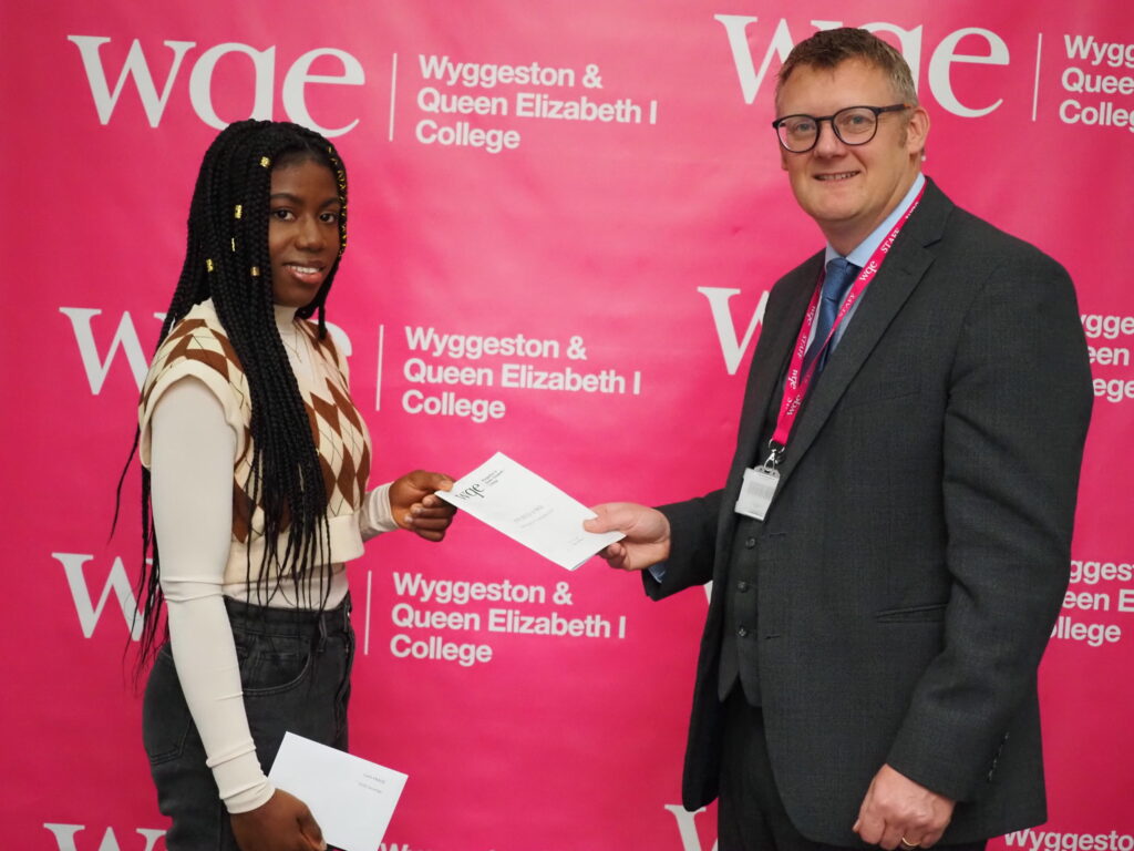 A student receives a certificate from a man in front of a Wyggeston & Queen Elizabeth I College banner.