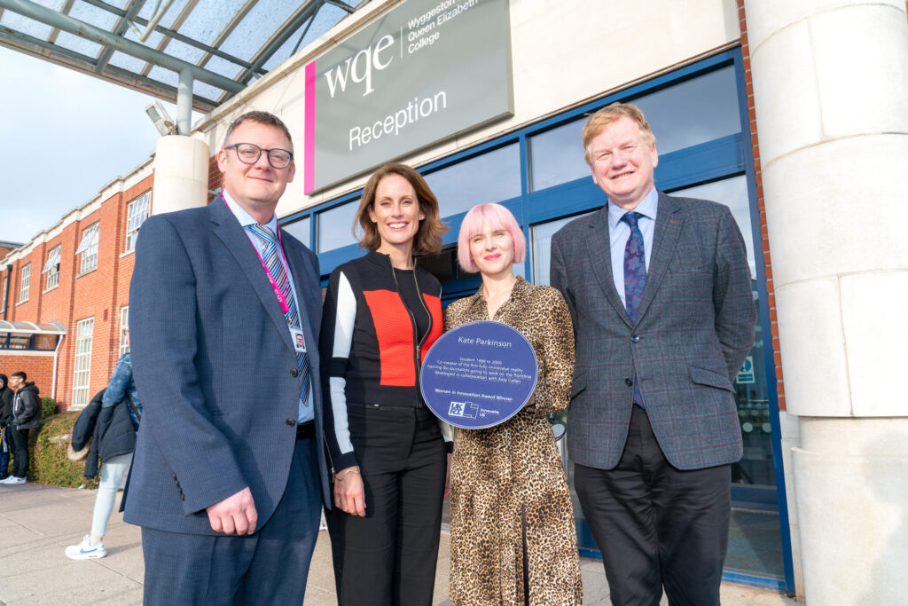 Four people stand smiling outside a building’s reception, one holding a blue plaque, with “wqe” signage above.
