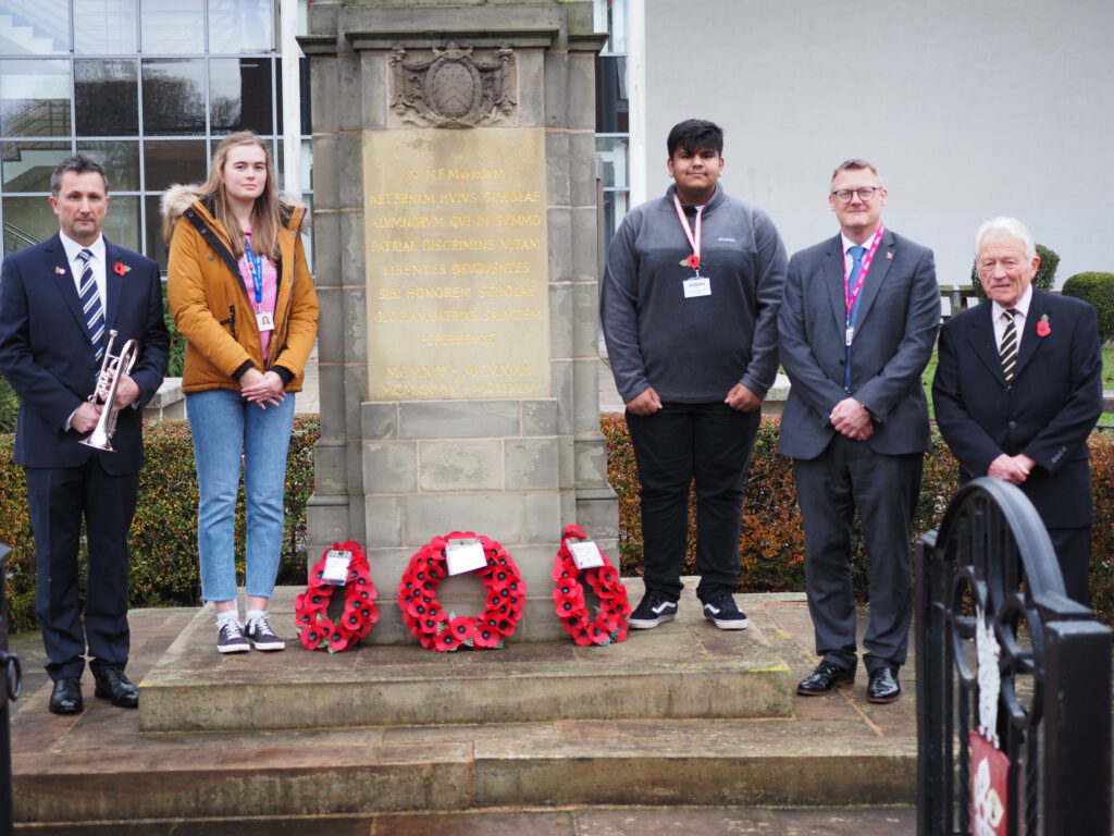Five people stand by a war memorial with wreaths of red poppies on the steps in front of the monument.