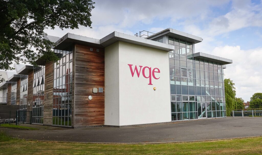 Modern building with large glass windows and wqe logo on a white wall, surrounded by grass and trees.