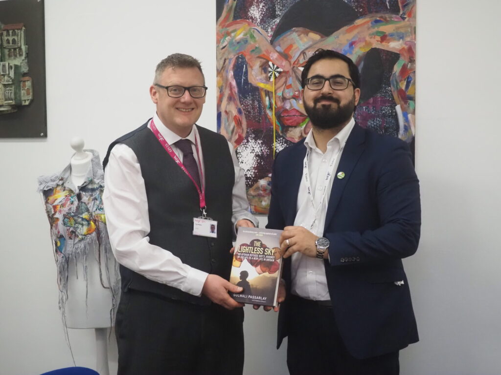 Two men in suits smiling, one handing a book titled The Limitless Sky to the other, art in background.
