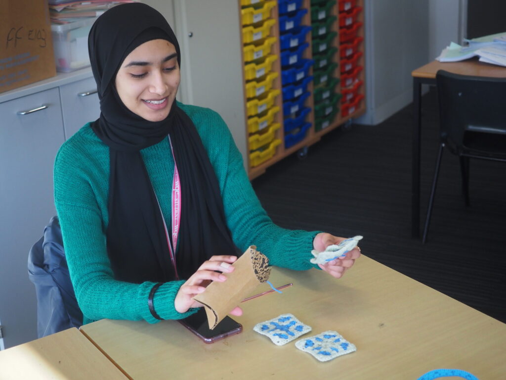 A woman in a green sweater smiles while making crafts at a table in a classroom.