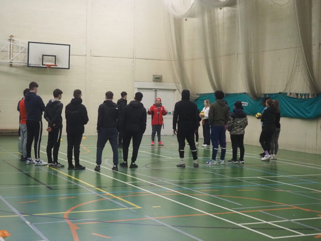 A group of people stand in a sports hall, listening to an instructor near a basketball hoop.