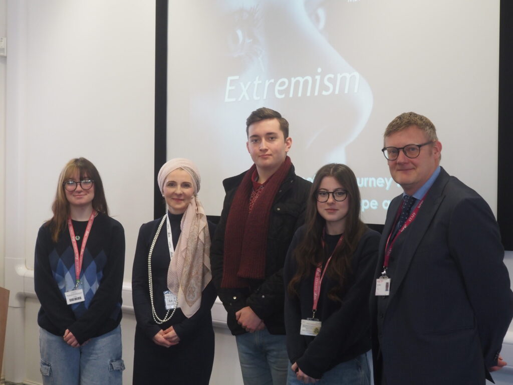 Five people stand in front of a screen displaying the word Extremism in a classroom setting.