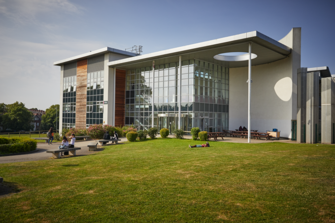Modern glass-fronted building with people sitting and relaxing on benches and grass in a sunny outdoor area.