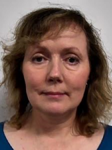 A woman with light brown, wavy hair and a neutral expression, wearing a blue top, against a plain background.