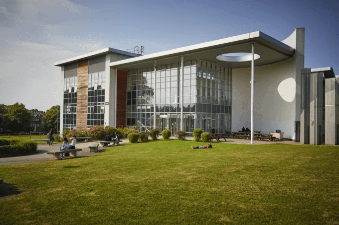 Modern glass-fronted building with people relaxing on benches and grass in front under a clear sky. For more information or inquiries, contact us.