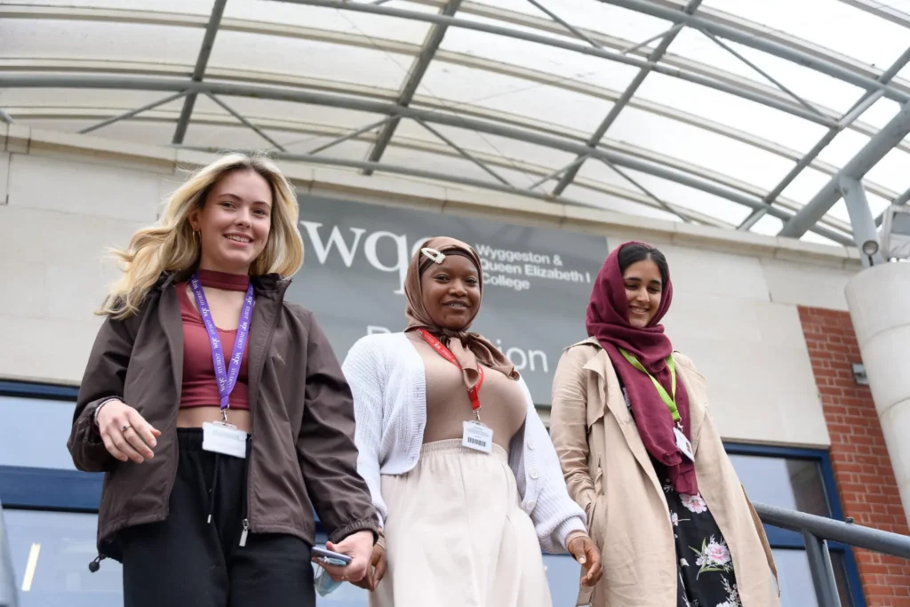 Three female students wearing lanyards and smiling as they walk down steps outside a college building.
