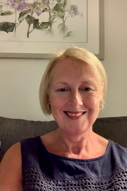 Smiling woman with short blonde hair sitting on a couch, with a floral painting on the wall behind her.