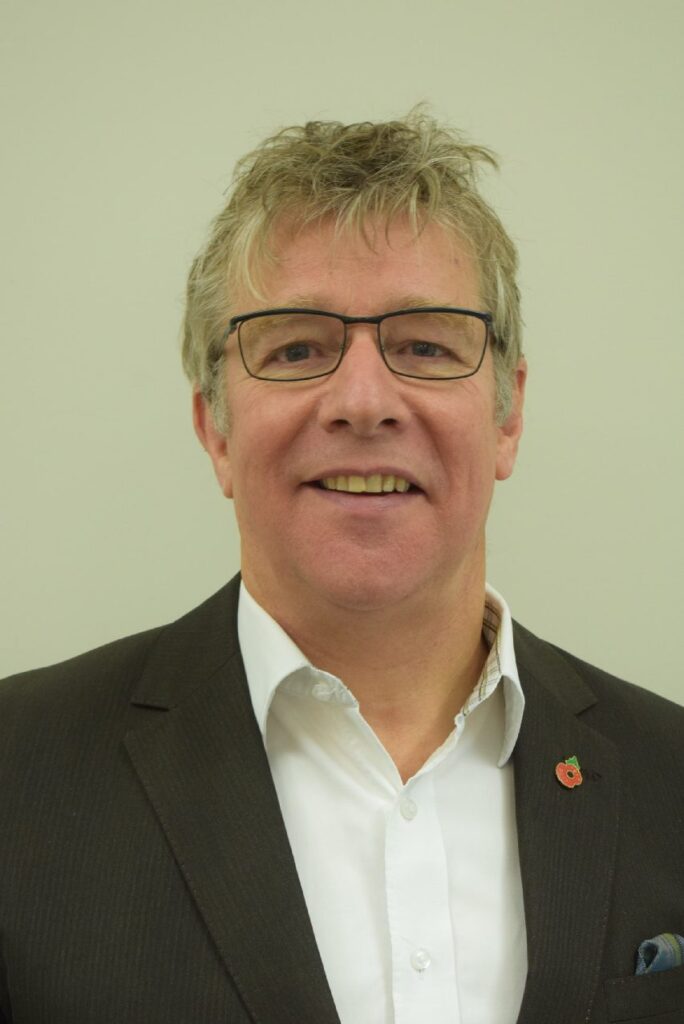 Man with glasses in a dark suit and white shirt, smiling against a plain light background.