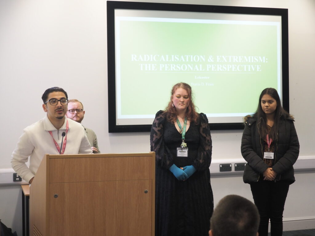 Four speakers stand at a podium in front of a screen reading “Radicalisation & Extremism: The Personal Perspective.”.