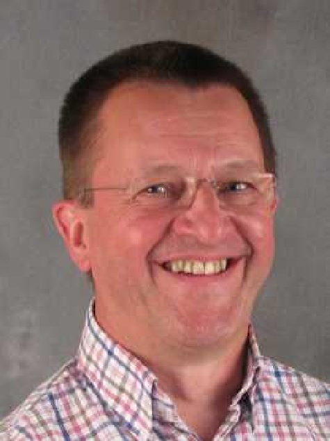 A man with glasses and a plaid shirt smiles in front of a plain gray background.