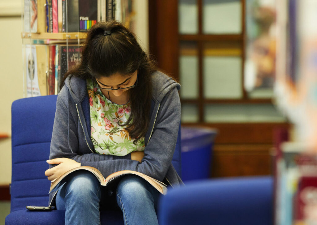 A young woman sits on a blue chair, reading a book with her arms crossed in a library, where safeguarding measures ensure a safe and welcoming environment.