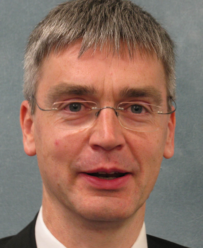 Man with short gray hair, glasses, and a suit, posing in front of a plain blue background.
