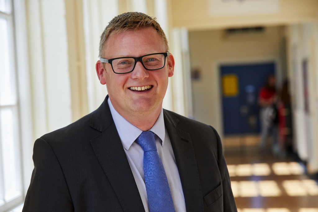 Smiling man in a suit and glasses stands in a brightly lit hallway with windows and blurred people in the background.