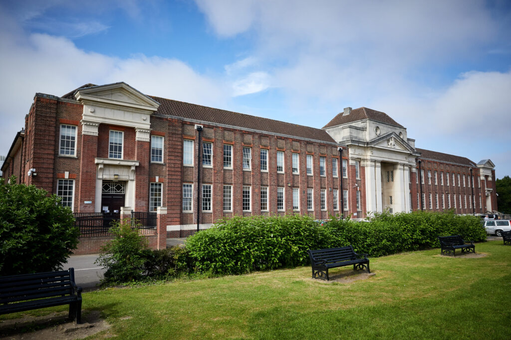 Large red-brick building with white columns, green bushes, and benches on a grassy lawn under a blue sky.