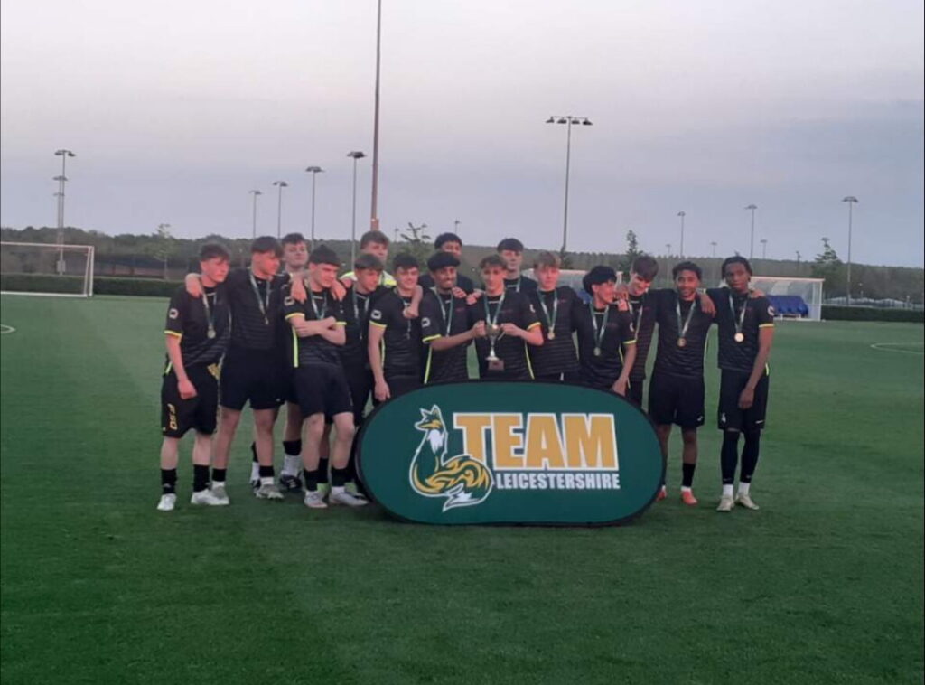 A photograph of WQE Men's Football First team huddled together whilst holding the Team Leicestershire Cup trophy.