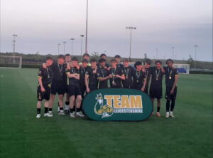 A photograph of WQE Men's Football First team huddled together whilst holding the Team Leicestershire Cup trophy.