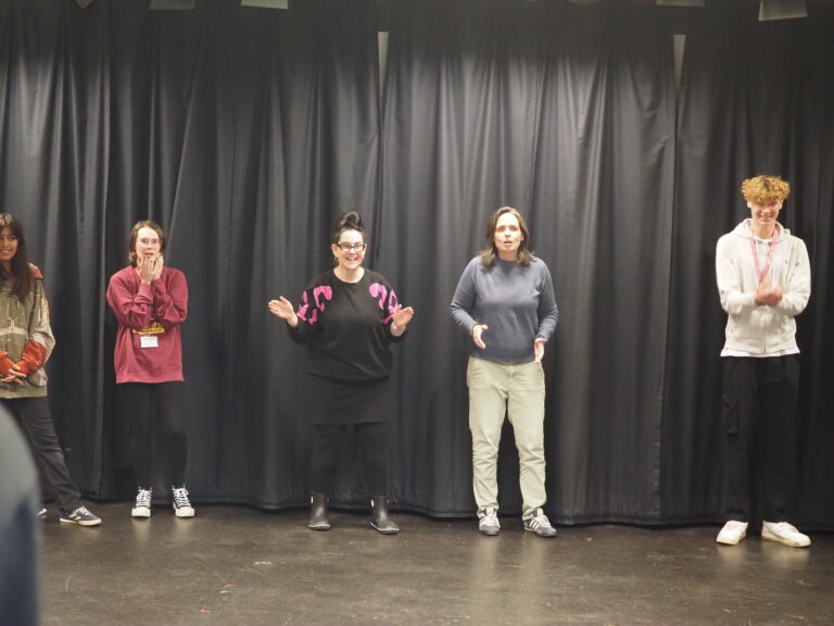 Five people stand spaced apart on a stage in front of black curtains, some clapping and smiling.