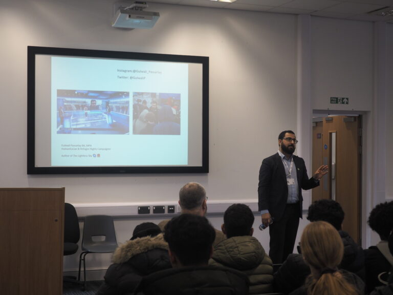 A man gives a presentation to an audience in a classroom, with slides projected on the wall behind him.