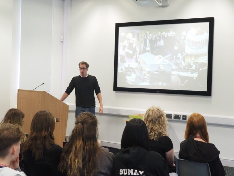 A man gives a presentation to a seated audience, with images displayed on a large screen behind him.