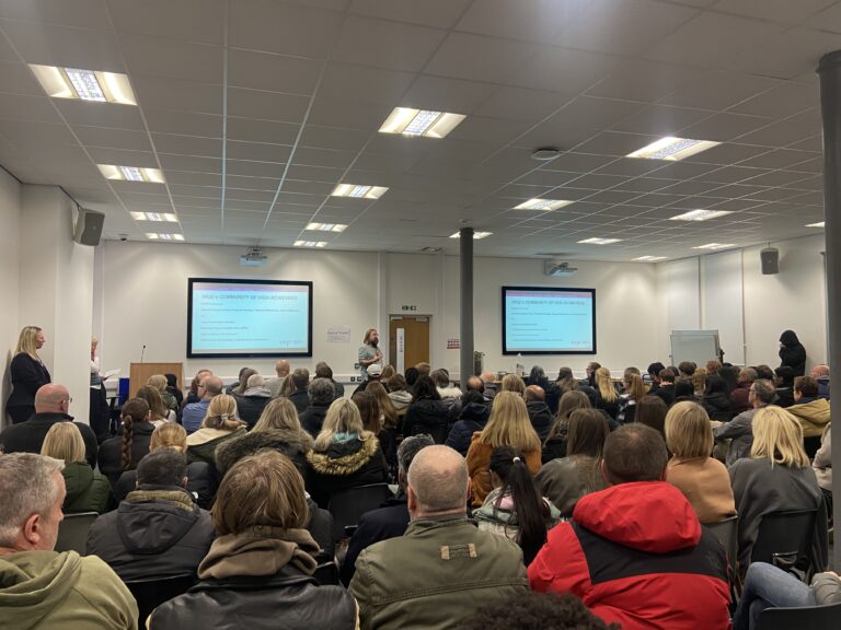 A large audience listens to a presenter in a bright conference room with slides displayed on screens.