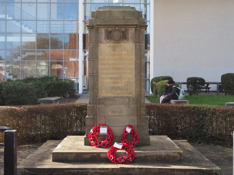 Stone war memorial with three red poppy wreaths at its base, set in front of a modern glass building.