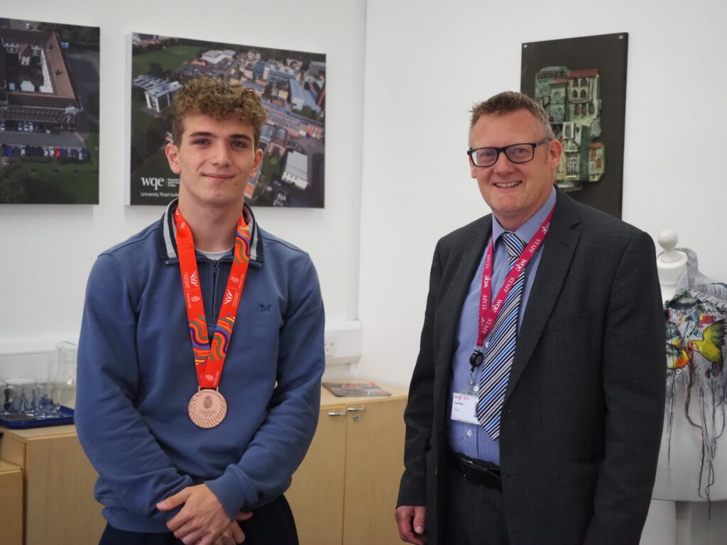 Young man wearing a medal stands next to a man in a suit; both are smiling in an office setting.