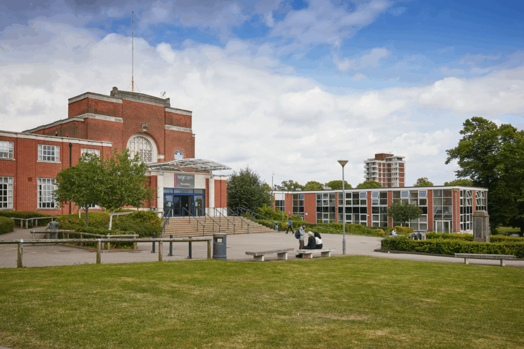 Red-brick university building with large windows, steps, and students sitting outside on benches under a blue sky.