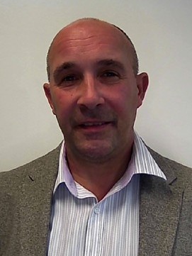 A man in a grey blazer and striped shirt smiles in front of a plain light background.