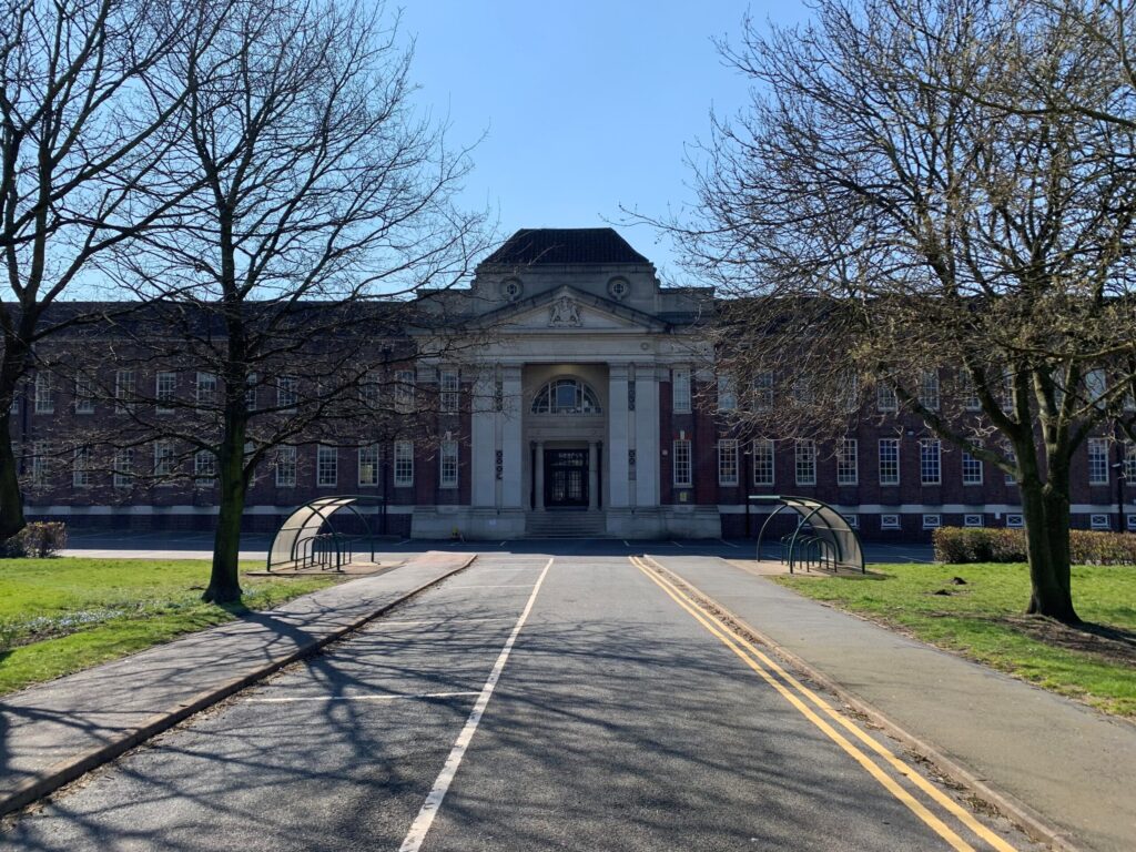 Large red-brick building with columns, seen from a tree-lined road on a sunny day.