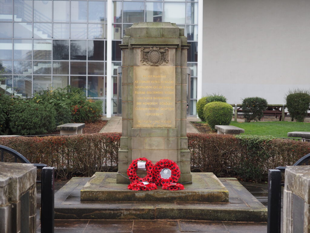 A stone war memorial with red poppy wreaths at its base, in a courtyard with bushes and a modern building behind.