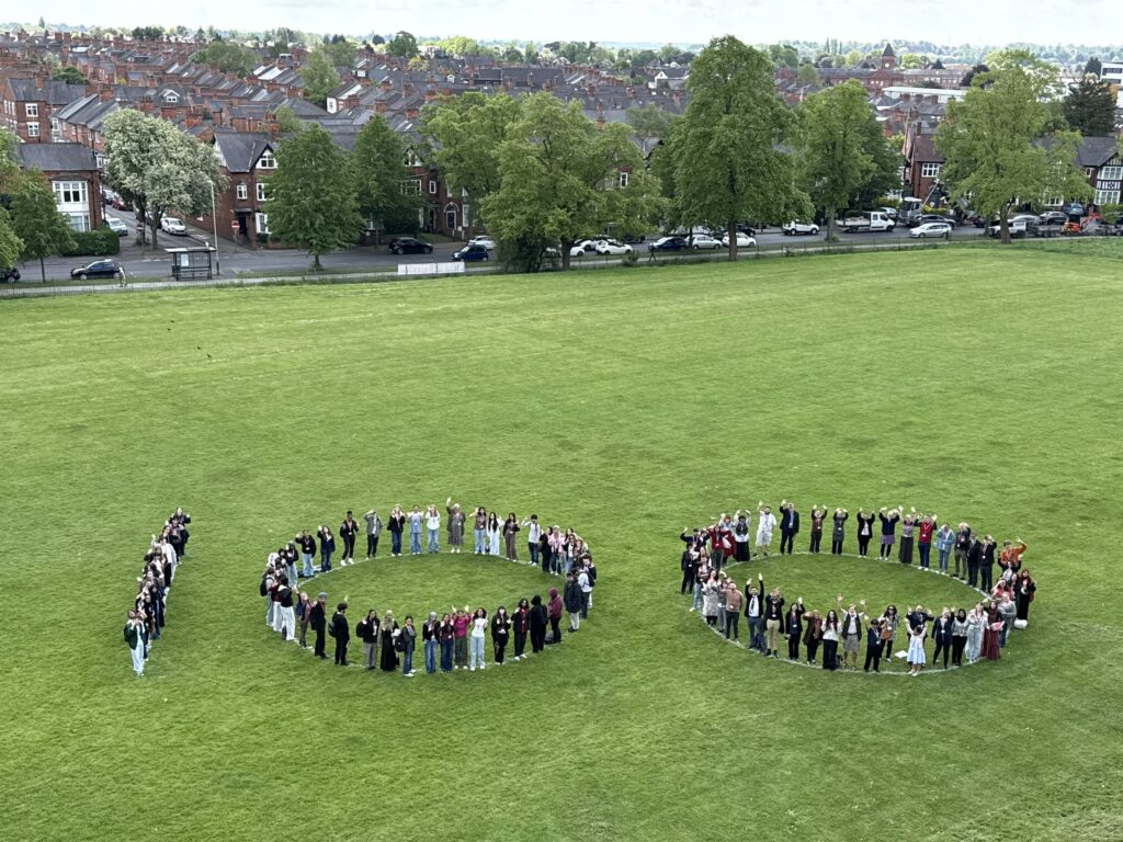 A large group of people stand in a field, forming the number 100 on the grass.