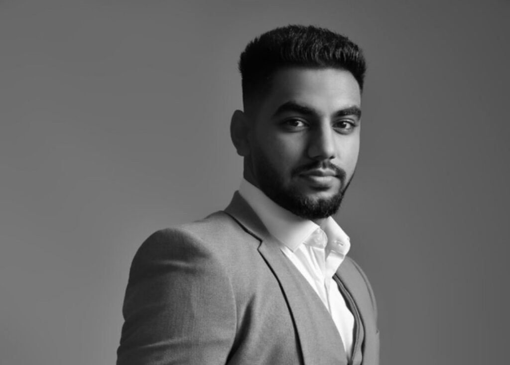 A man in a suit poses confidently against a plain, neutral background in a black-and-white photo.