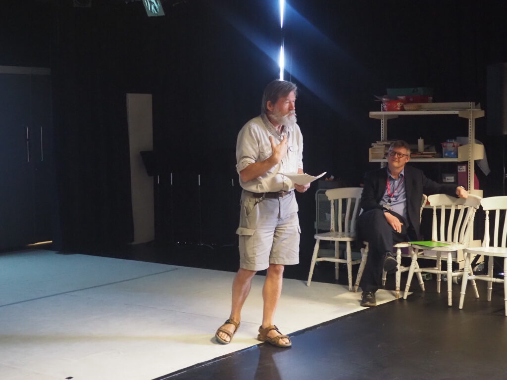 A man in shorts and sandals speaks onstage whilst another person sits on a white chair in the background.