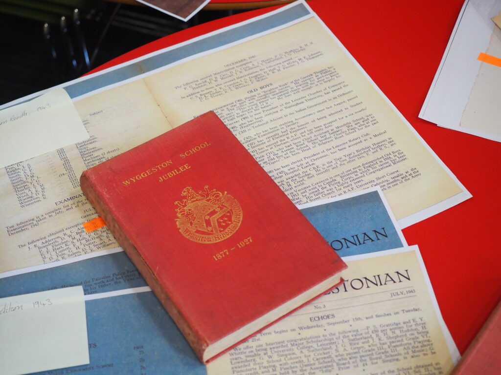 A red Wyggeston School Jubilee book lies on top of various old school documents and papers.