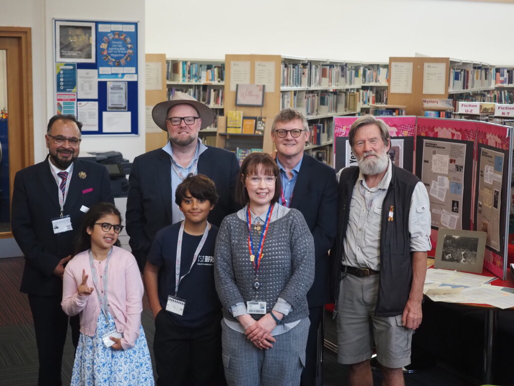 A group of adults and children pose and smile together in a library with display boards behind them.