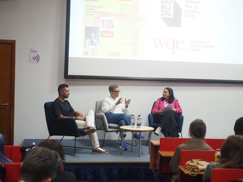 Three people sit on stage in discussion, with a presentation projected behind them and an audience watching.