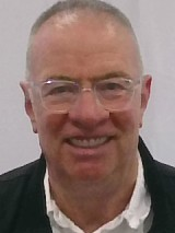 Smiling older man with glasses, short grey hair, wearing a white shirt and dark jacket against a light background.