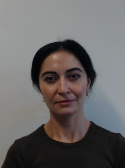 Woman with dark hair and brown shirt looking at the camera, standing against a plain light background.
