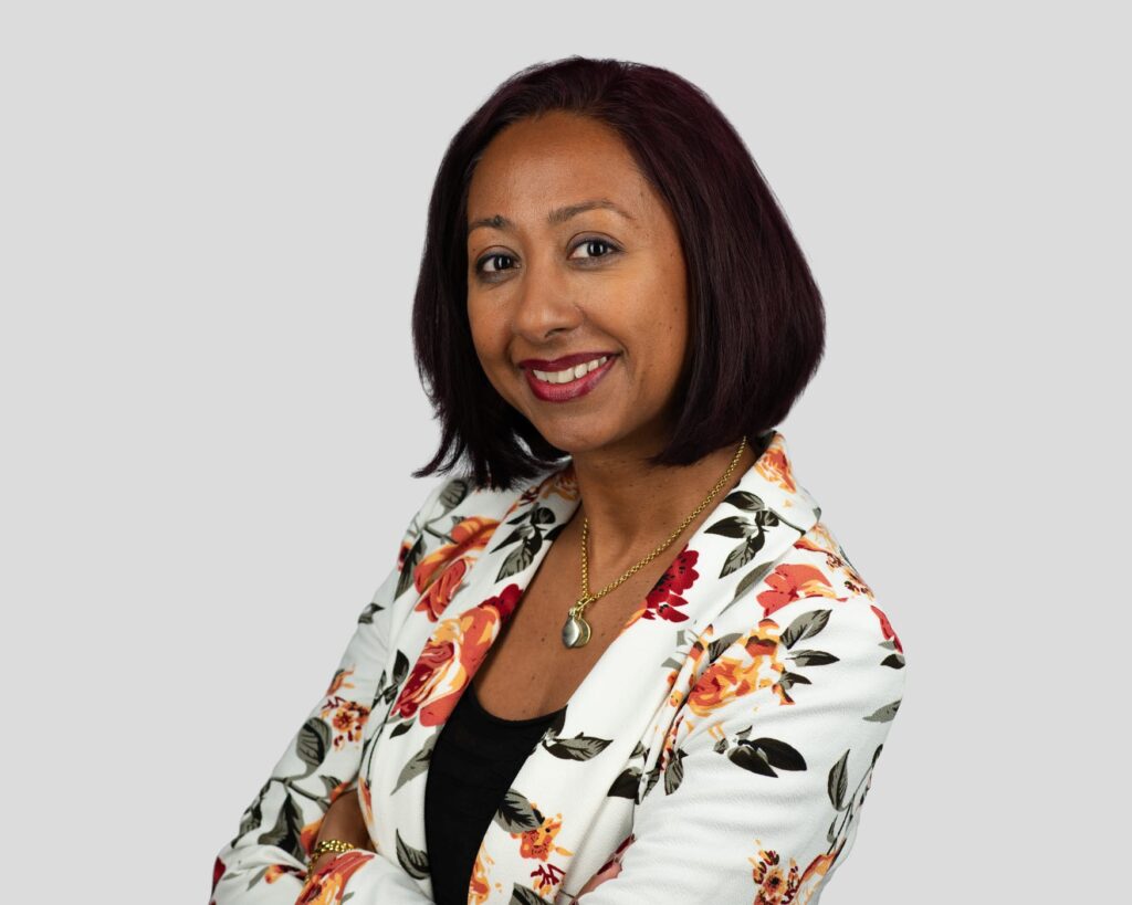 Woman smiling with arms crossed, wearing a floral blazer, black top, necklace, on a plain light background.